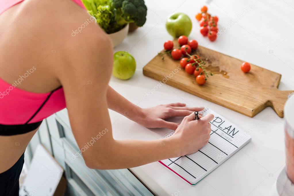 cropped view of sportswoman writing in notebook with plan lettering in kitchen 