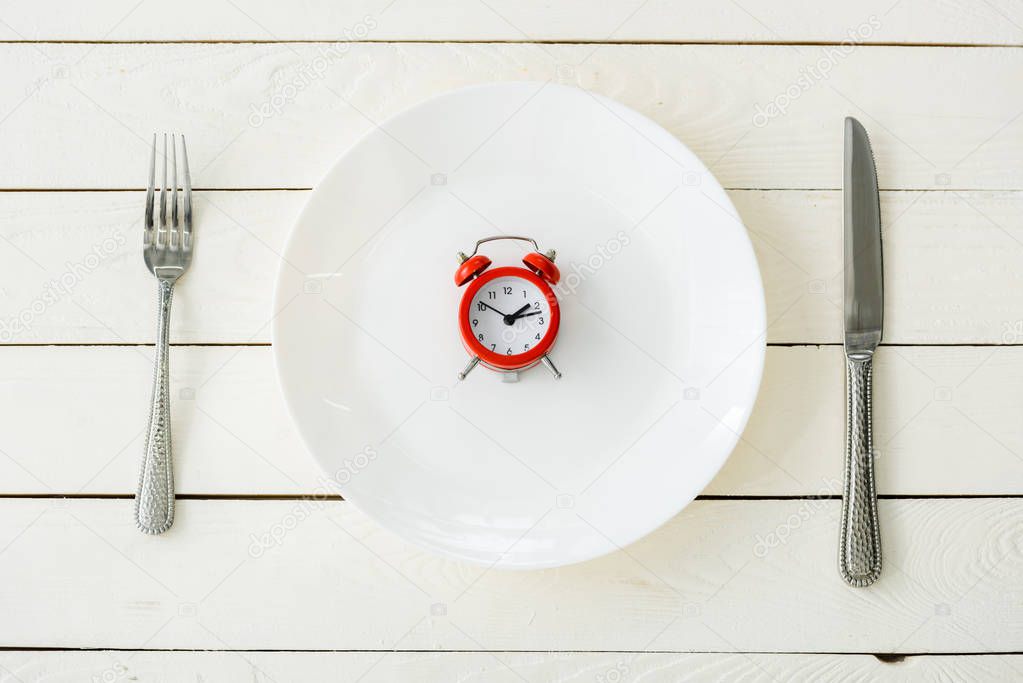 top view of white plate with red alarm clock near cutlery on wooden surface 