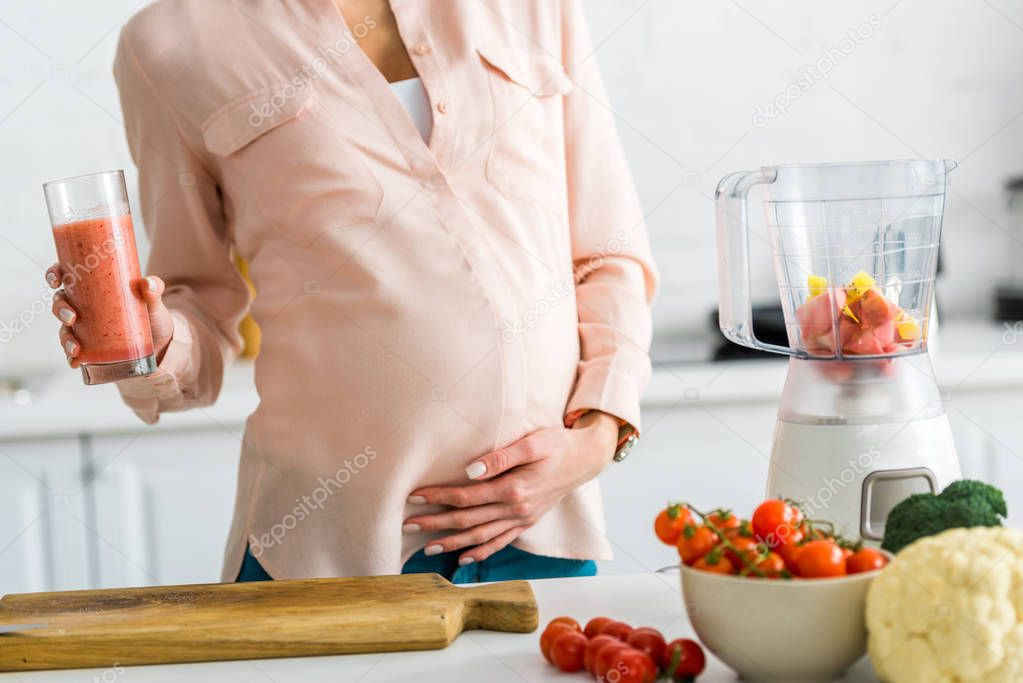 cropped view of pregnant woman holding glass of smoothie near vegetables in kitchen 