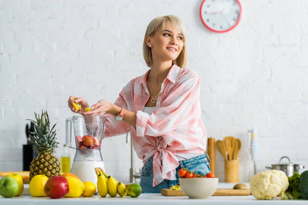  attractive girl adding ingredients in blender while smiling in kitchen 