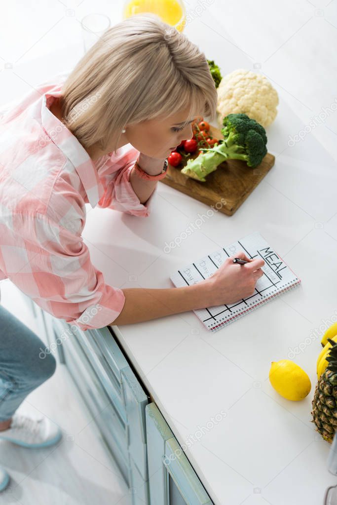 overhead view of blonde girl writing in notebook with meal lettering near ingredients in kitchen