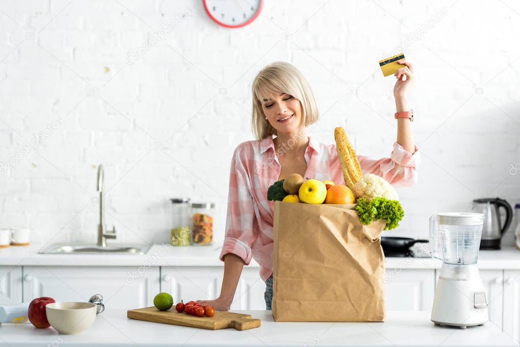 happy blonde girl holding credit card near paper bag with groceries 