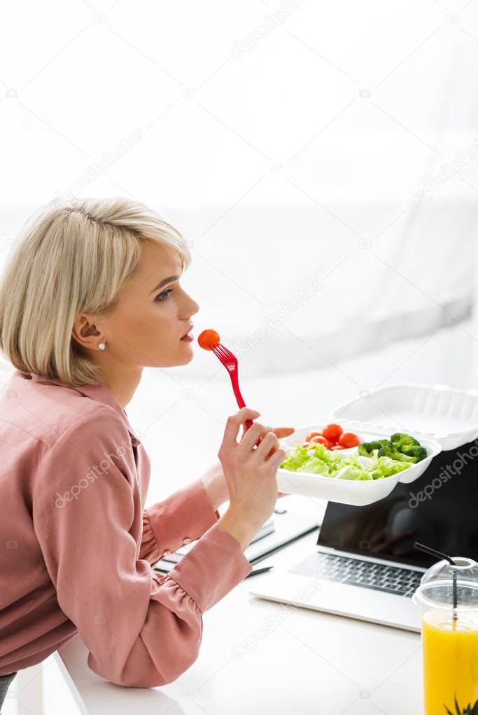 blonde freelancer sitting near laptop and eating cherry tomato  