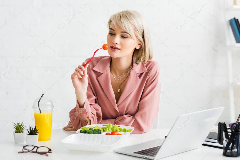 blonde freelancer holding fork with cherry tomato near laptop