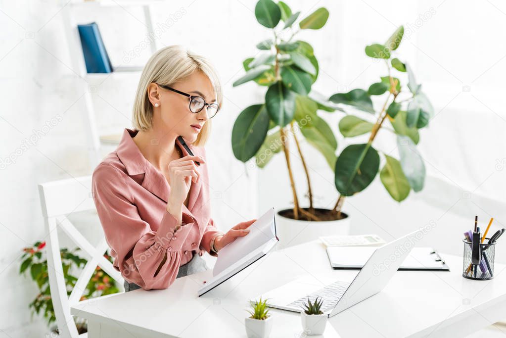 pensive young blonde freelancer in glasses holding pen and notebook while sitting near laptop 