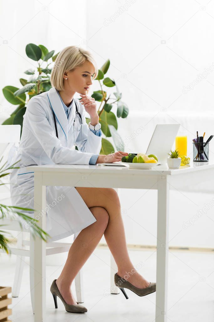 blonde nutritionist using laptop near plate with organic food