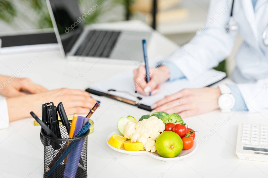 cropped view of nutritionist writing diagnosis near patient and plate with organic food 