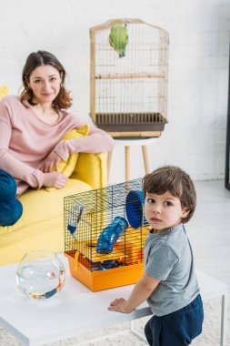 balık kase ve pet kafes ile masa yakın ayakta sevimli çocuk seçici odak ve sarı kanepe üzerinde dinlenmek gülümseyen anne
