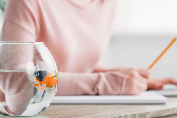 partial view of woman sitting at table near aquarium with gold fish