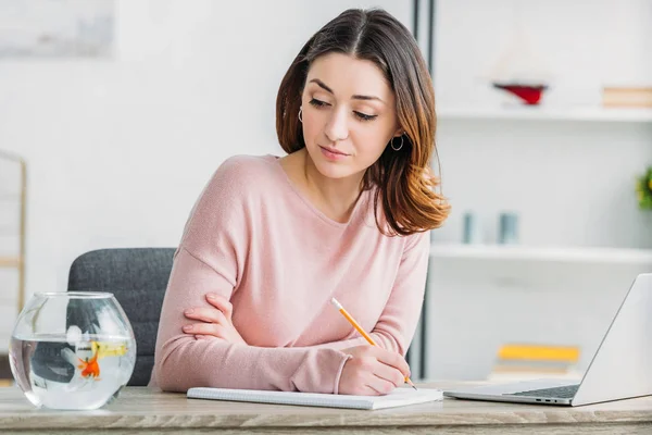 Mooie Vrouw Holding Potlood Kijken Naar Fishbowl Zittend Aan Tafel — Stockfoto