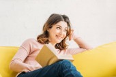 usmívající se atraktivní žena s knihou na žluté pohovce doma