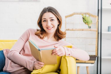 attractive woman holding book and looking at camera at home clipart