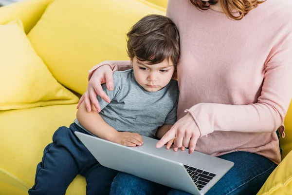 自宅で黄色のソファに座っている間にノートパソコンを使用して 愛らしい息子と女性のクロップビュー  — 無料ストックフォト