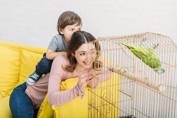 smiling mother and son looking at green parrot in bird cage at home