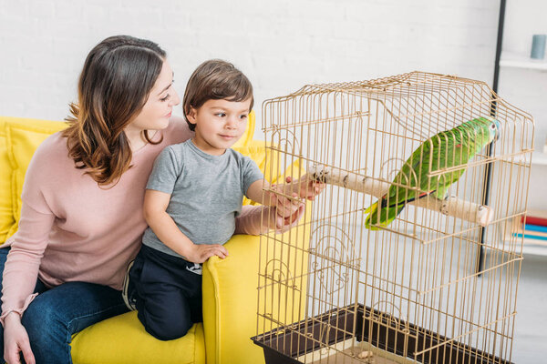 happy mother with adorable son looking at green parrot in bird cage at home
