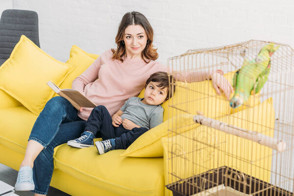 smiling mother with adorable son sitting on sofa near bird age with green parrot