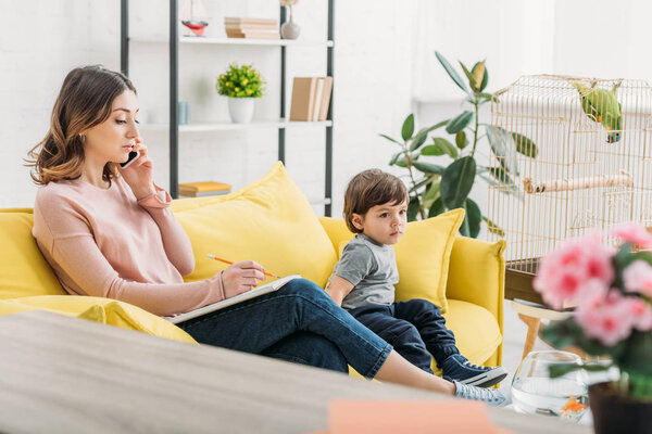 serious woman talking on smartphone while sitting on sofa near adorable son