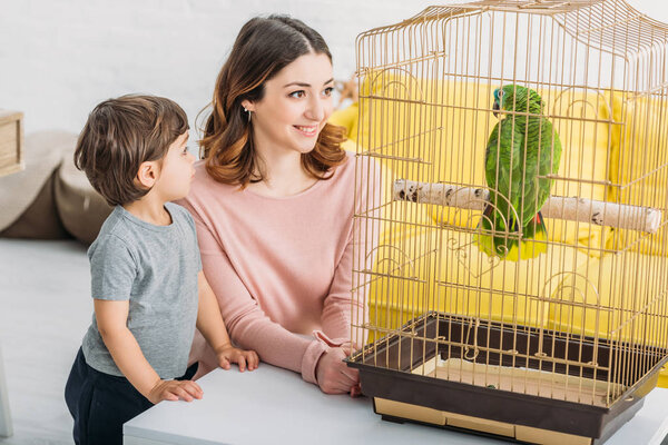 smiling attractive woman with adorable son looking at green parrot in bird cage