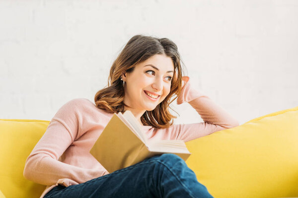smiling attractive woman with book resting on yellow sofa at home