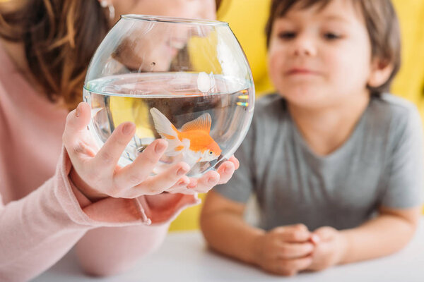 partial view of smiling woman holding fish bowl with bright gold fish near cute son