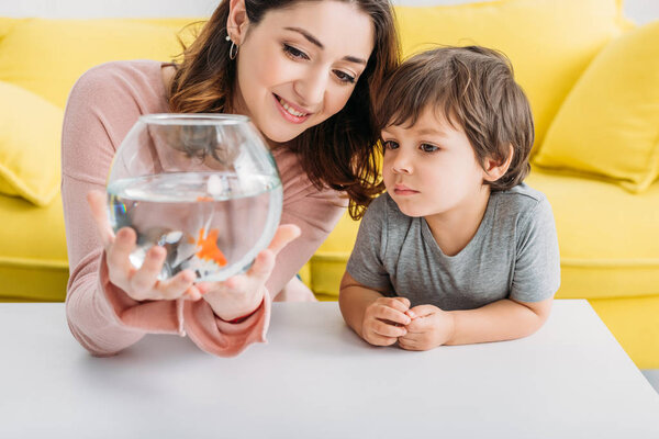 smiling mother holding fish bowl near adorable son at home
