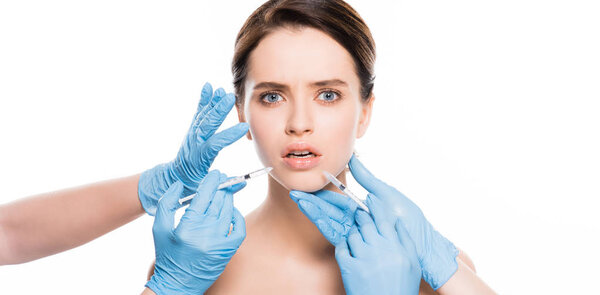 Панорамный снимок косметологов в латексных перчатках, касающихся лица потрясенной девушки, держащей в руках изолированные на белом шприцы

