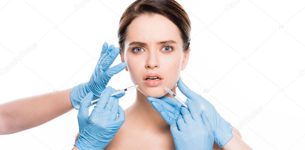 panoramic shot of cosmetologists in latex gloves touching face of shocked girl while holding  syringes isolated on white