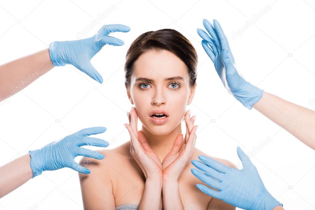 cropped view of plastic surgeons in blue latex gloves near face of surprised girl isolated on white