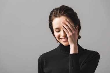 cheerful young woman in black jumper covering eye isolated on grey  clipart
