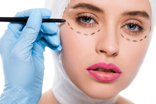 крупным пластмассовым хирургом в латексной перчатке, держащим маркер рядом с женщиной с отметинами на лице, изолированными на белом
 