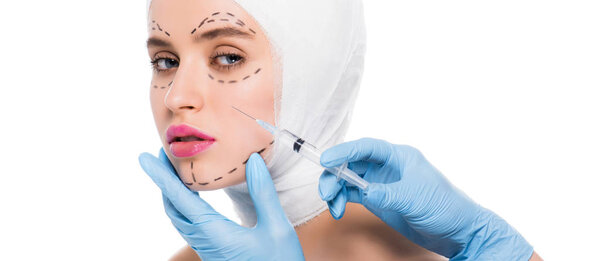 Панорамный снимок пластического хирурга в латексных перчатках, держащего шприц возле лица молодой женщины с отметинами, изолированными на белом
 