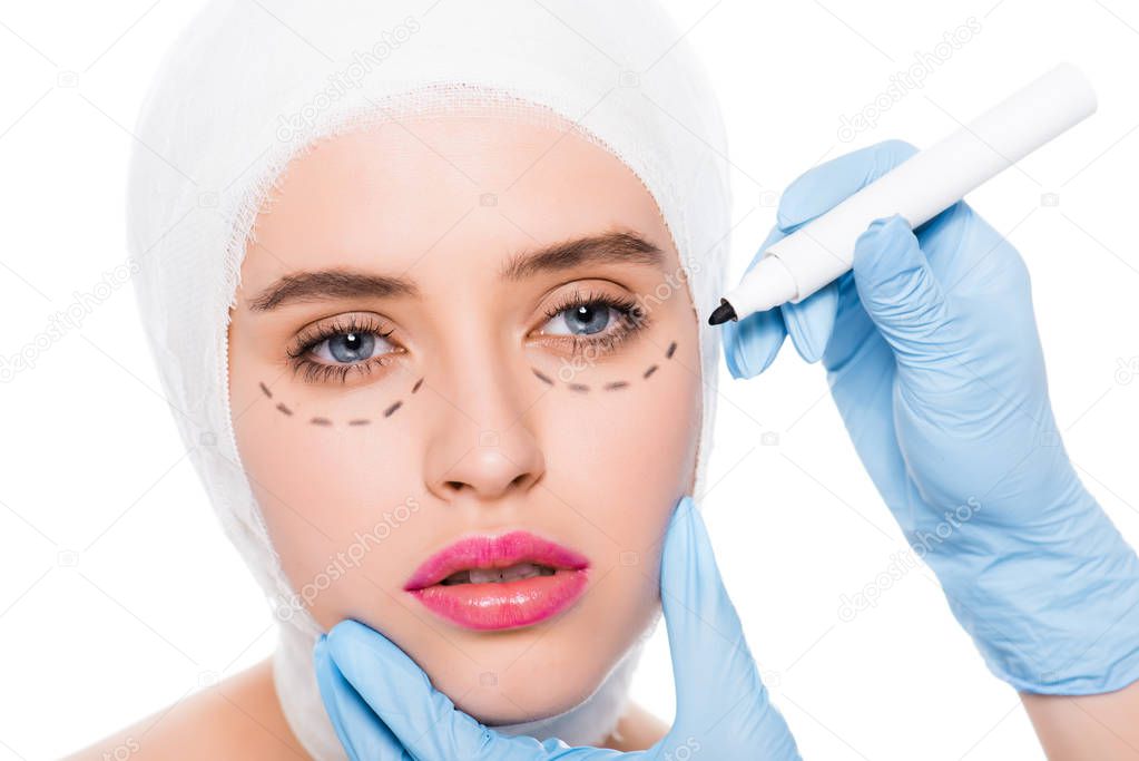 cropped view of plastic surgeon in latex gloves holding marker pen near woman with marks on face isolated on white 