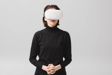 brunette young woman wearing virtual reality headset while standing with clenched hands isolated on grey 