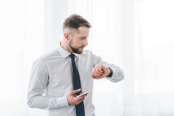 handsome bearded man in suit holding smartphone while looking at watch