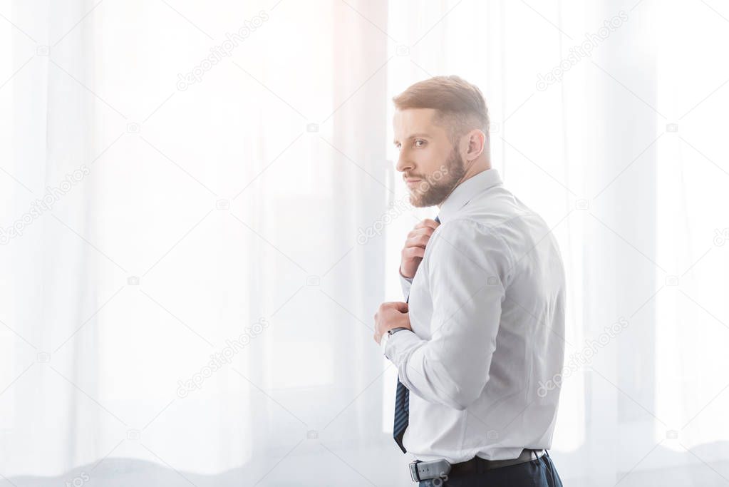 handsome bearded man touching tie while standing at home