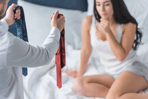 ベッドの中でペンシブブルネットの女性の近くに赤と青のネクタイを保持している男のトリミングビュー — ストック写真