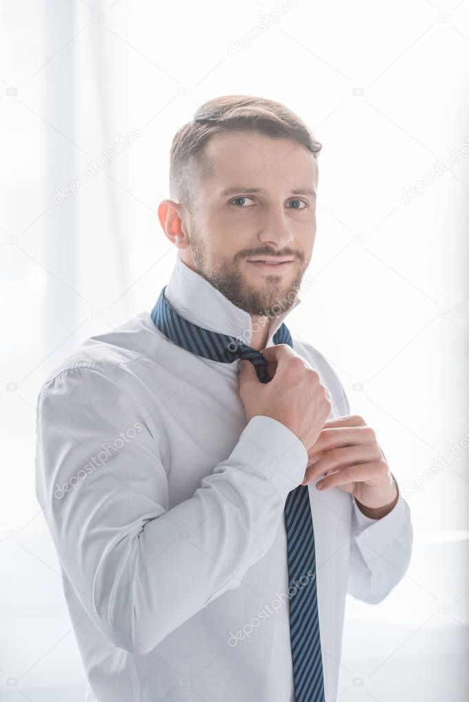happy bearded man in formal wear smiling while touching tie 