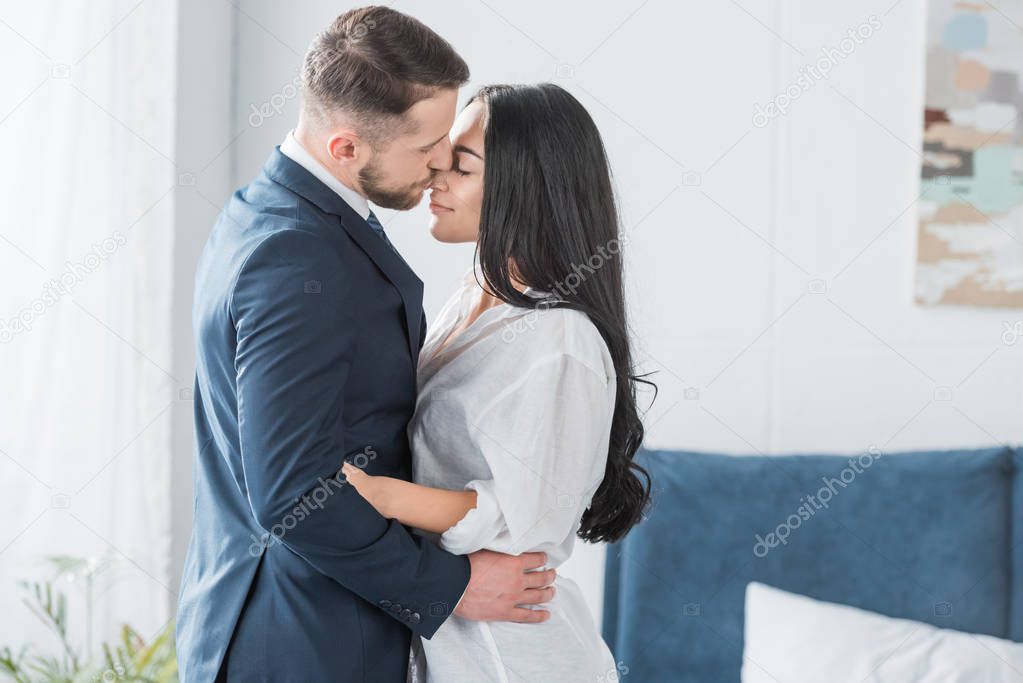 happy bearded man in suit kissing nose of attractive girlfriend in white shirt 