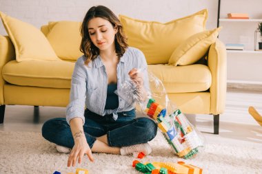 Tired woman in jeans sitting on carpet with toy blocks clipart