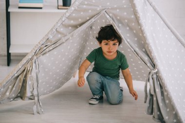 cute kid in jeans sitting in grey wigwam at home clipart
