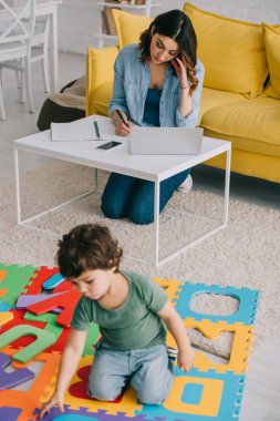 child playing with puzzle mat while mother working wih laptop clipart