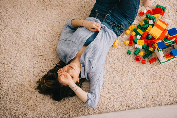 overheaf view of tired woman lying on carpet with colorful toy blocks