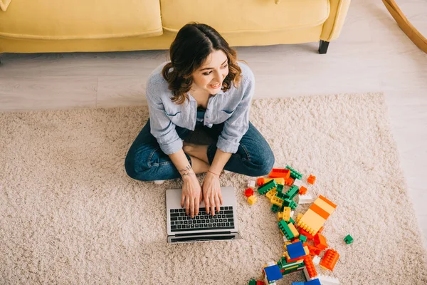 おもちゃのブロックの近くにカーペットの上に座って ラップトップを使用して笑顔の女性のオーバーヘッドビュー — ストック写真