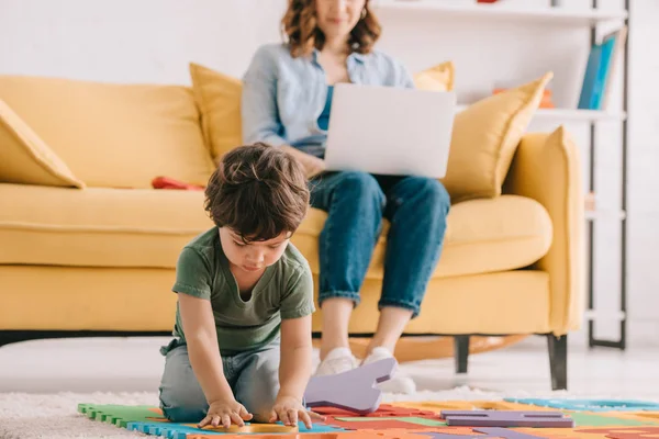 ラップトップを使用して母親ながらパズルマットで遊ぶ緑のTシャツのかわいい子供 — ストック写真