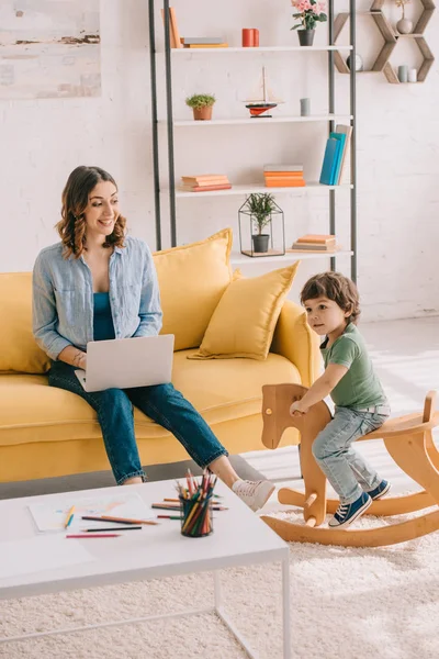 Kid Sitter Gungande Häst Medan Mamma Arbetar Med Laptop Vardagsrummet — Stockfoto