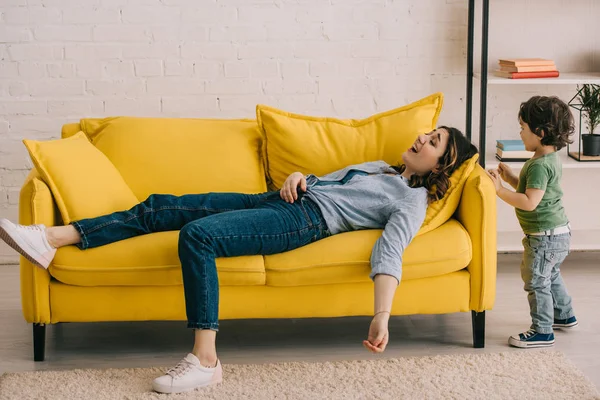 Little boy standing near tired mother lying on yellow sofa in living room — Stock Image Little Boy Standing Tired Mother Lying Yellow Sofa Living Room Royalty Free Stock Photos