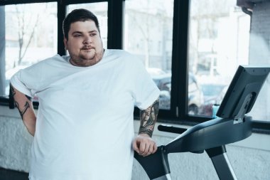 kilolu dövmeli adam spor merkezinde koşu bandı yakınında uzağa bakıyor