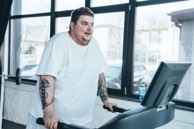 overweight tattooed man in white t-shirt running on treadmill at sports center clipart
