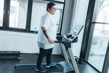spor salonunda koşu bandı üzerinde çalışan beyaz t-shirt kilolu dövmeli adam