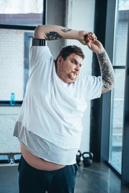 overweight tattooed man in white t-shirt stretching at sports center clipart